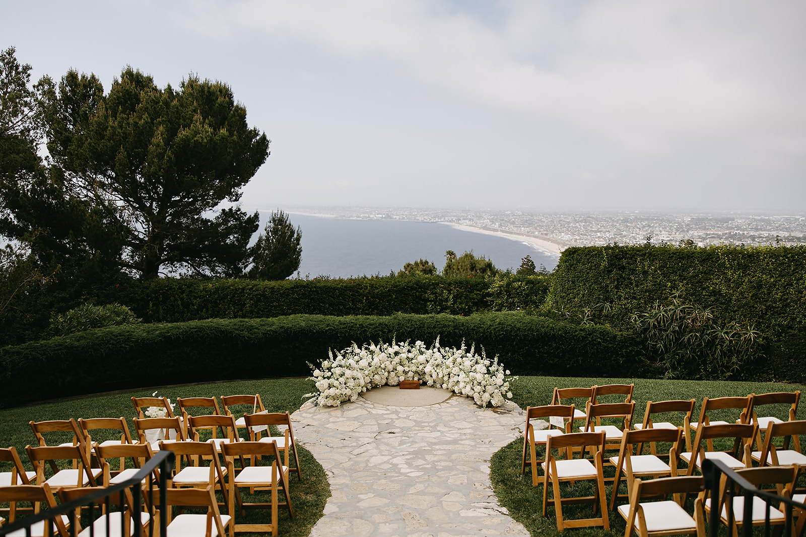 An outdoor wedding ceremony setup with wooden chairs arranged in rows facing an altar adorned with white flowers, overlooking a scenic ocean view under a clear sky at La Venta Inn