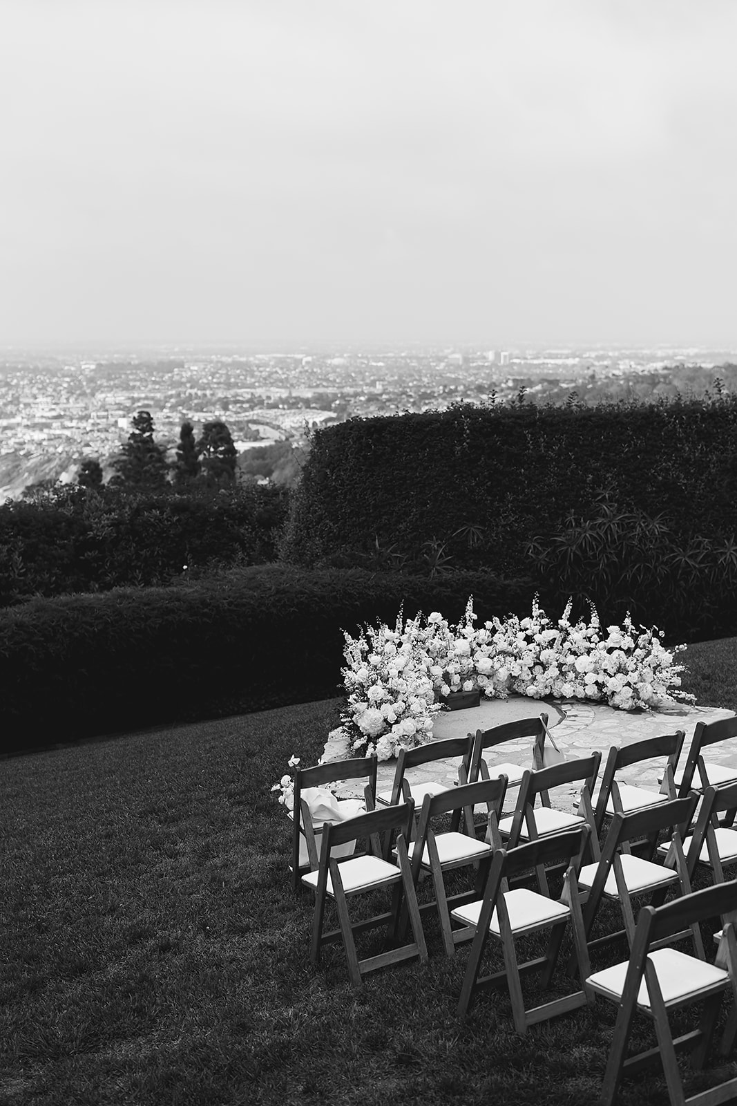 Rows of wooden chairs face a flower-adorned table outdoors, overlooking a cityscape under a cloudy sky.