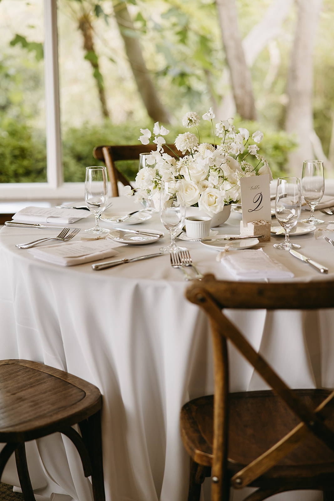 Elegant dining table set for an event with a white tablecloth, floral arrangements, and wooden chairs in a room with large windows and a view of greenery outside.