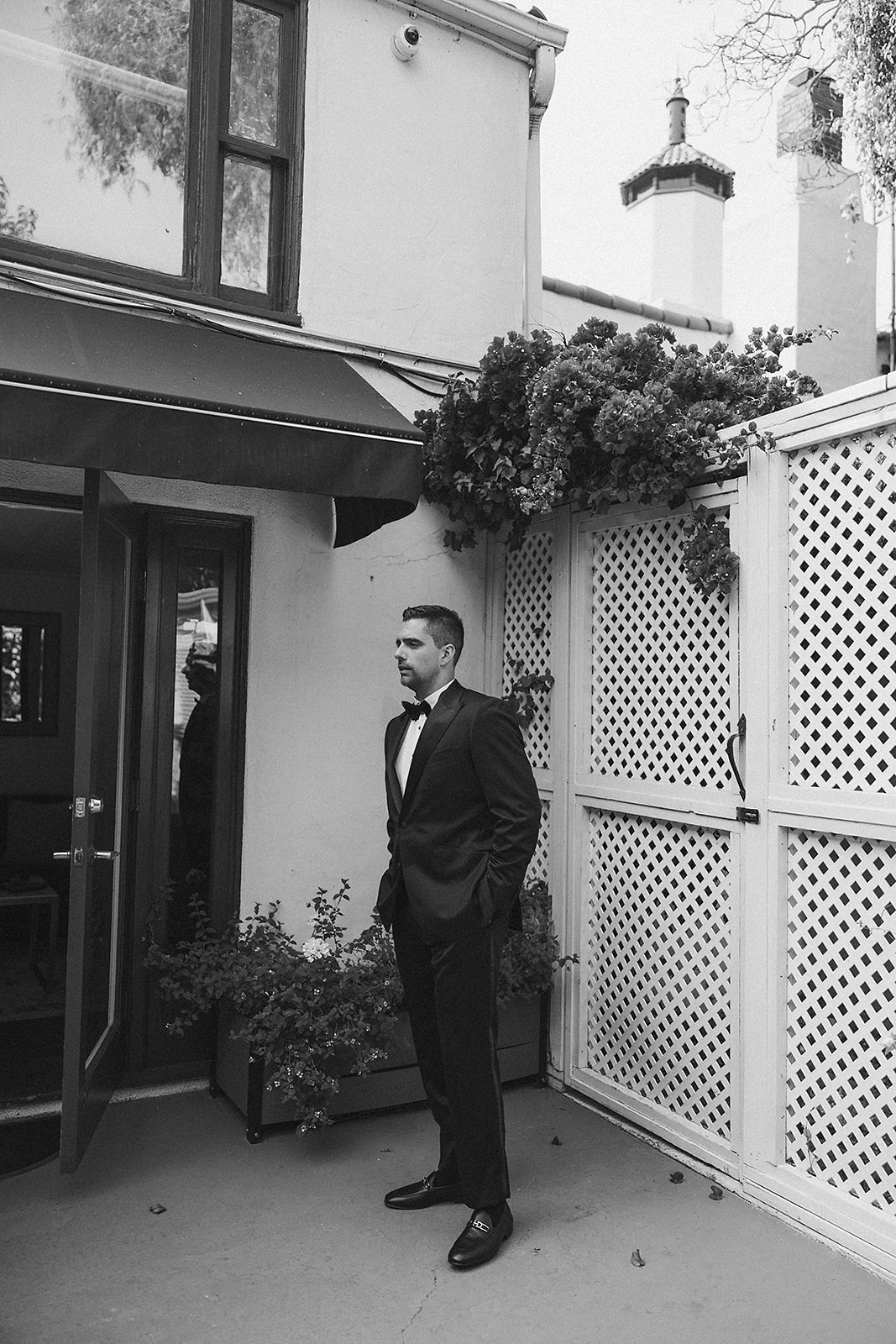 Two men in suits stand outside near a wall with flowers. One man adjusts the other's tie.