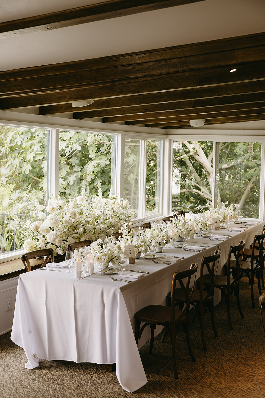 Elegant dining table set for an event with a white tablecloth, floral arrangements, and wooden chairs in a room with large windows and a view of greenery outside.