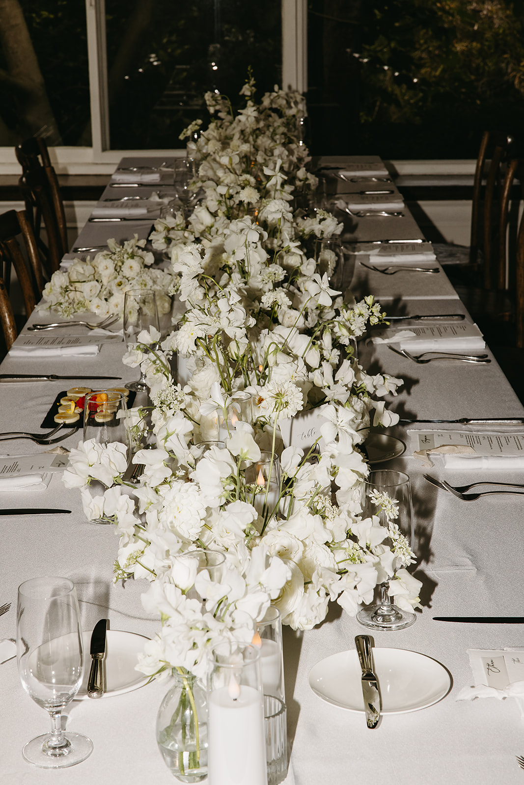 Elegant dining table with white floral arrangements, set with plates, glasses, cutlery, and a small platter of appetizers in the background.