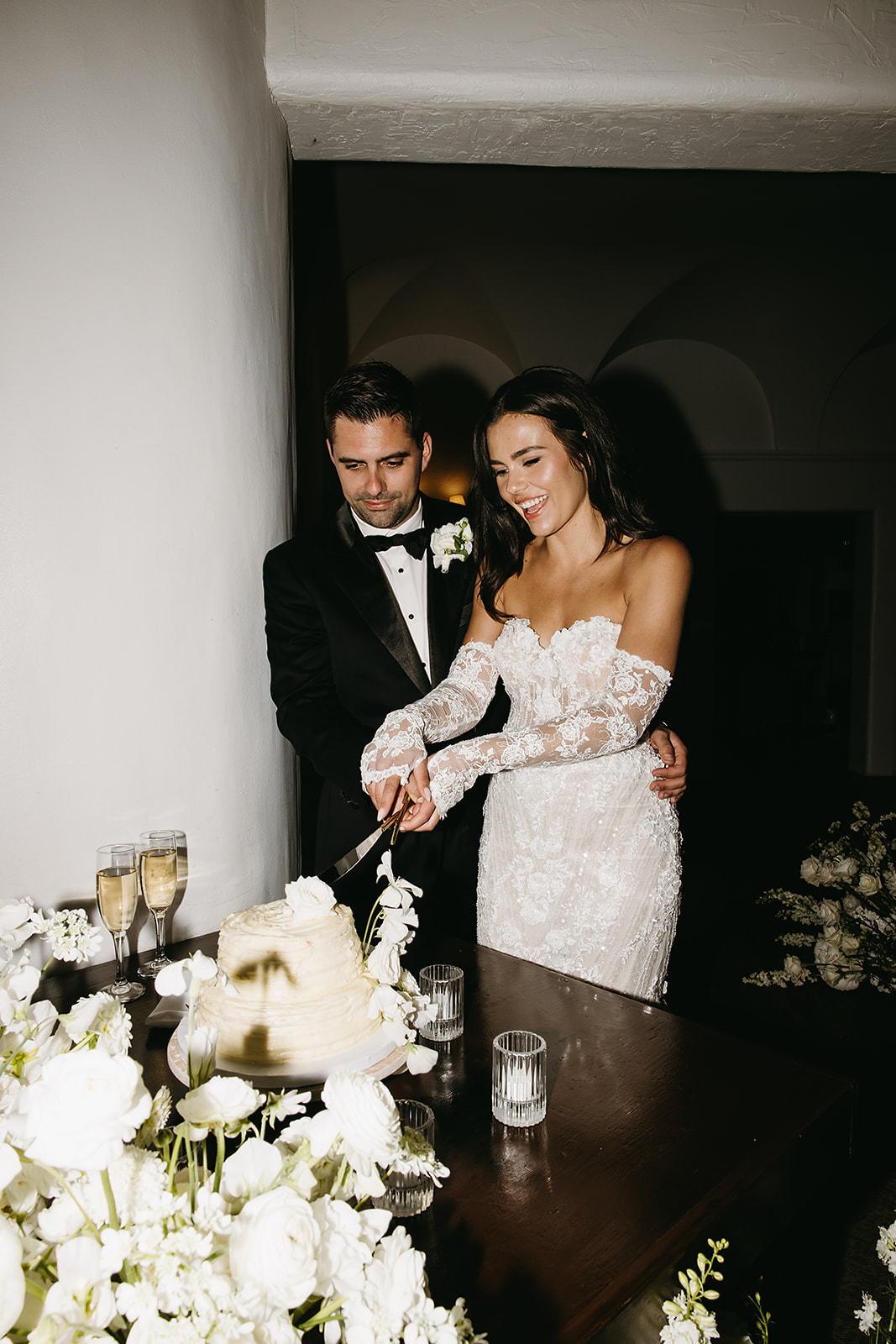 A bride and groom in formal attire cut a wedding cake together on a table adorned with white flowers and candles.