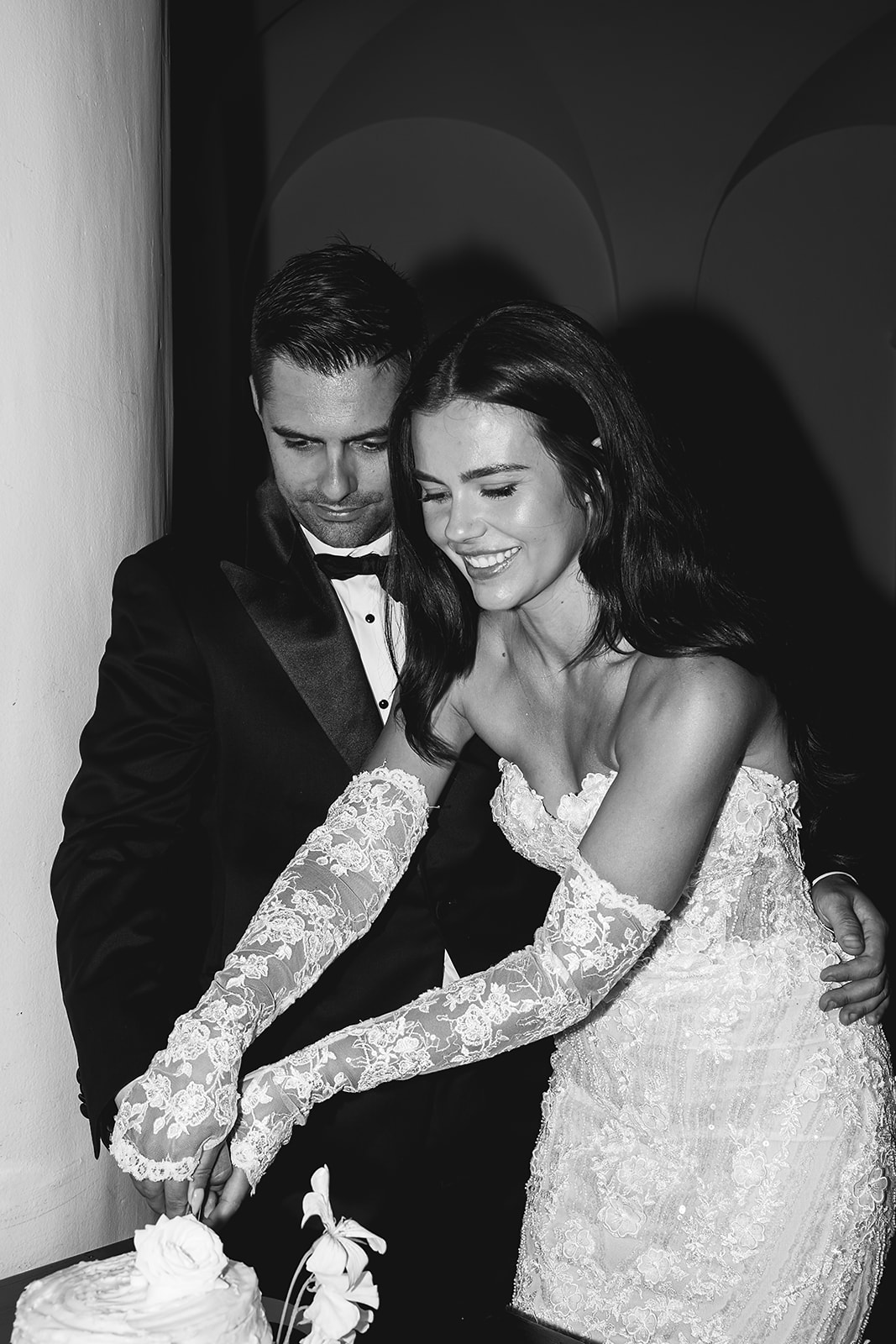 A bride and groom in formal attire cut a wedding cake together on a table adorned with white flowers and candles.