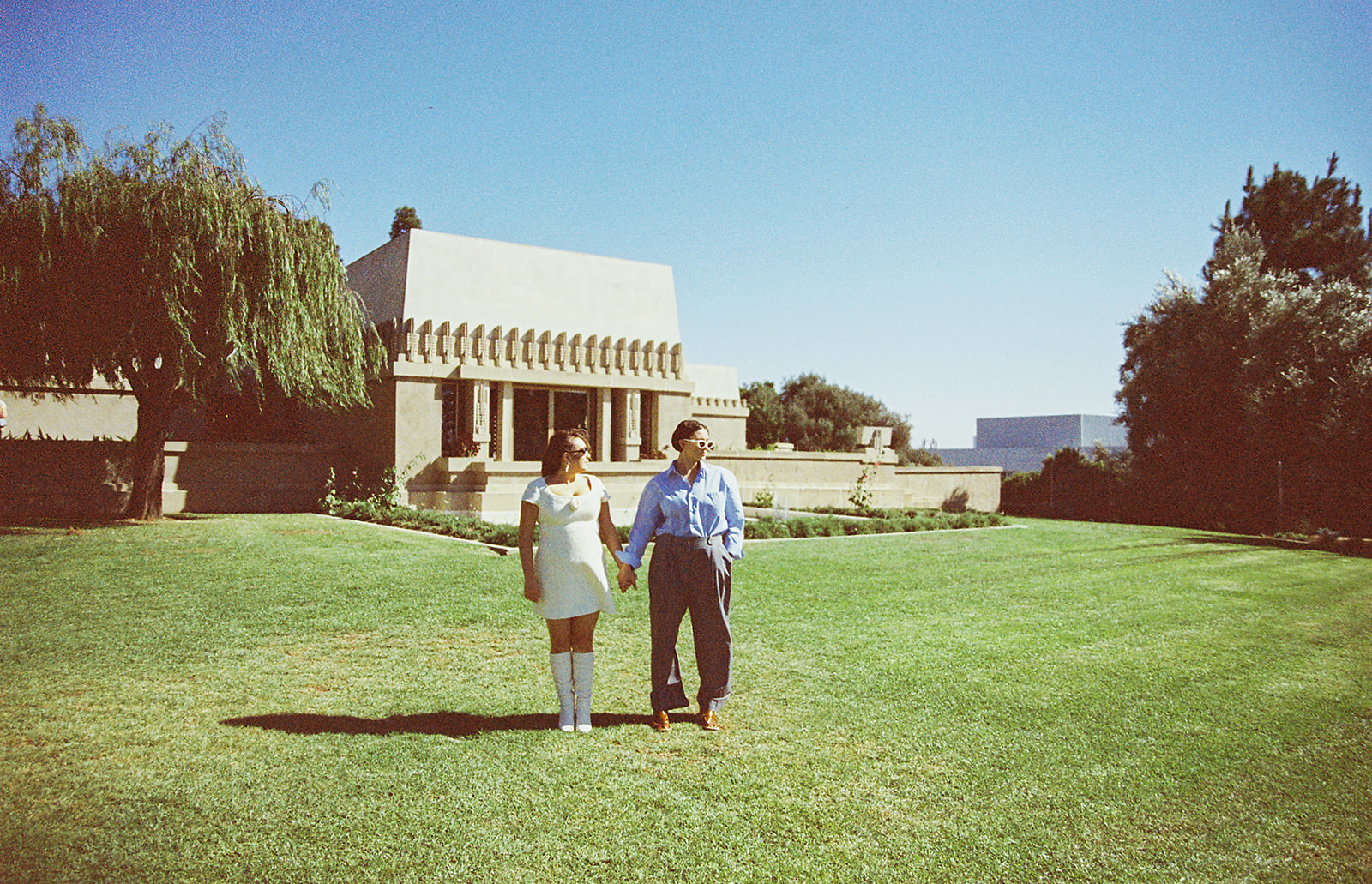 A couple holding hands on a sunny day with a cityscape in the background. wearing a blue shirt and gray pants, and the woman is in a white dress and boots.