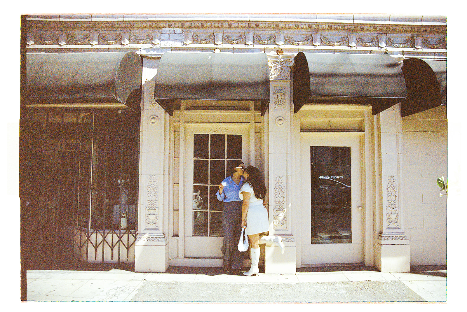 A person in a white dress and boots embraces another wearing a blue shirt and gray pants, standing outside a building with glass doors for their romantic engagement photos in LA