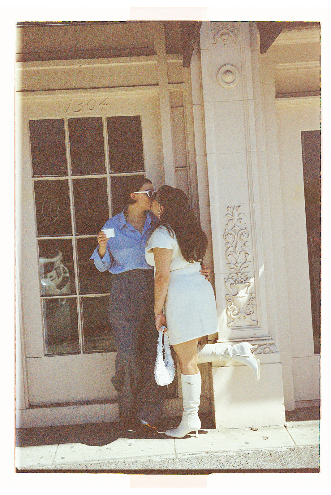 Two people share a kiss in front of a windowed building. One person wears a blue shirt and pants; the other wears a white dress and boots.