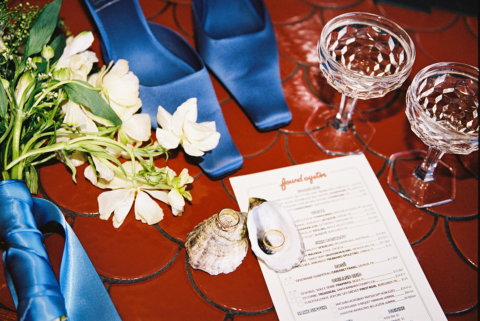 A pair of blue shoes, a bouquet, two crystal glasses, a menu, and seashells are arranged on a red-tiled surface.