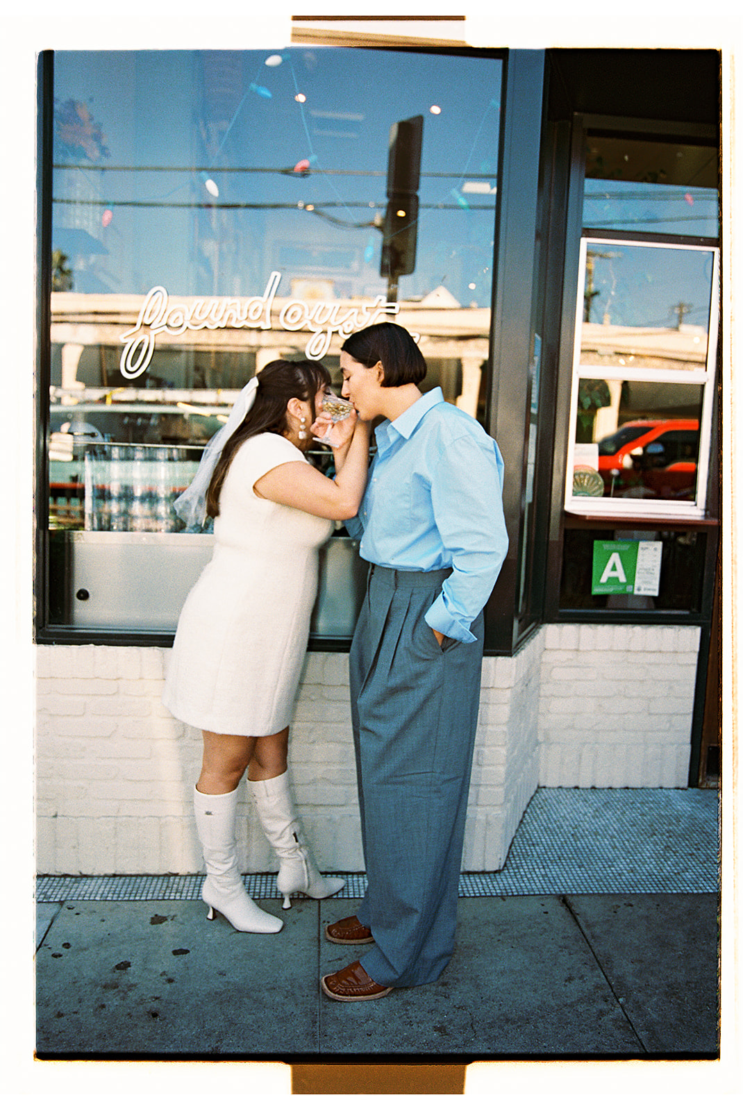 Two people in vintage dresses share a kiss outside a storefront with glass doors. One wears white boots and the other a long white dress for their romantic engagement photos in LA