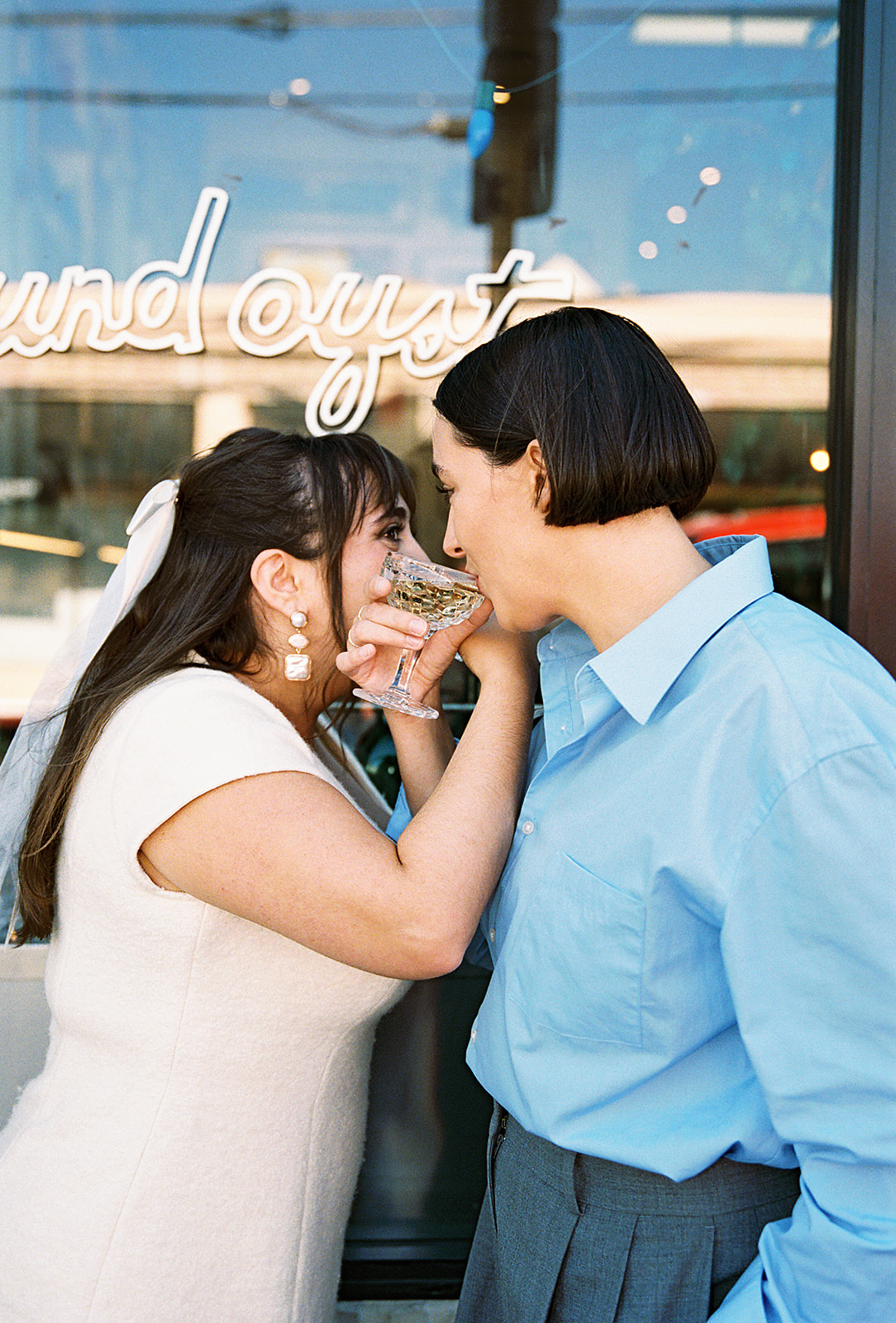 Two people in vintage dresses share a kiss outside a storefront with glass doors. One wears white boots and the other a long white dress for their romantic engagement photos in LA