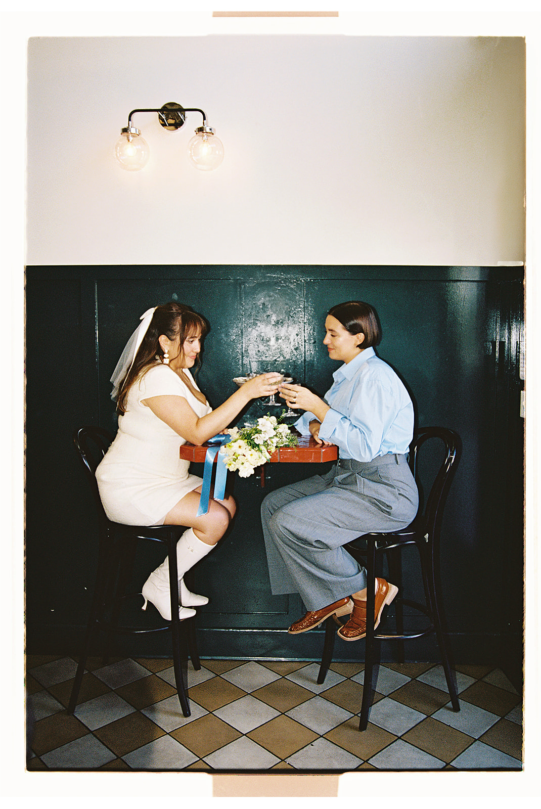  Two people sit at a small table in a cafe. One is dressed in a short white dress holding a bouquet, and the other wears a blue shirt and gray pants. They are holding hands and smiling.