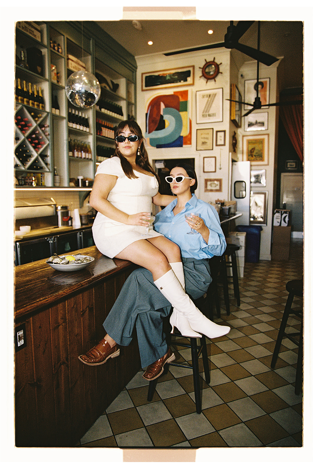 Two people in stylish outfits pose confidently on bar stools in a trendy, art-filled space. One person wears a white dress and boots, the other a blue shirt and pants. Both are wearing sunglasses.