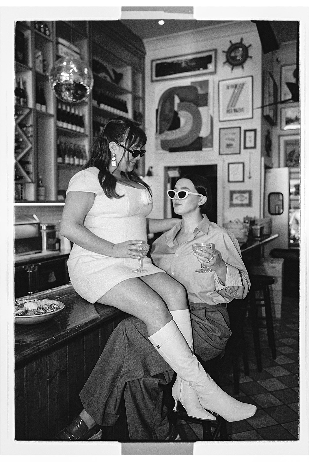 Two people in stylish outfits pose confidently on bar stools in a trendy, art-filled space. One person wears a white dress and boots, the other a blue shirt and pants. Both are wearing sunglasses.