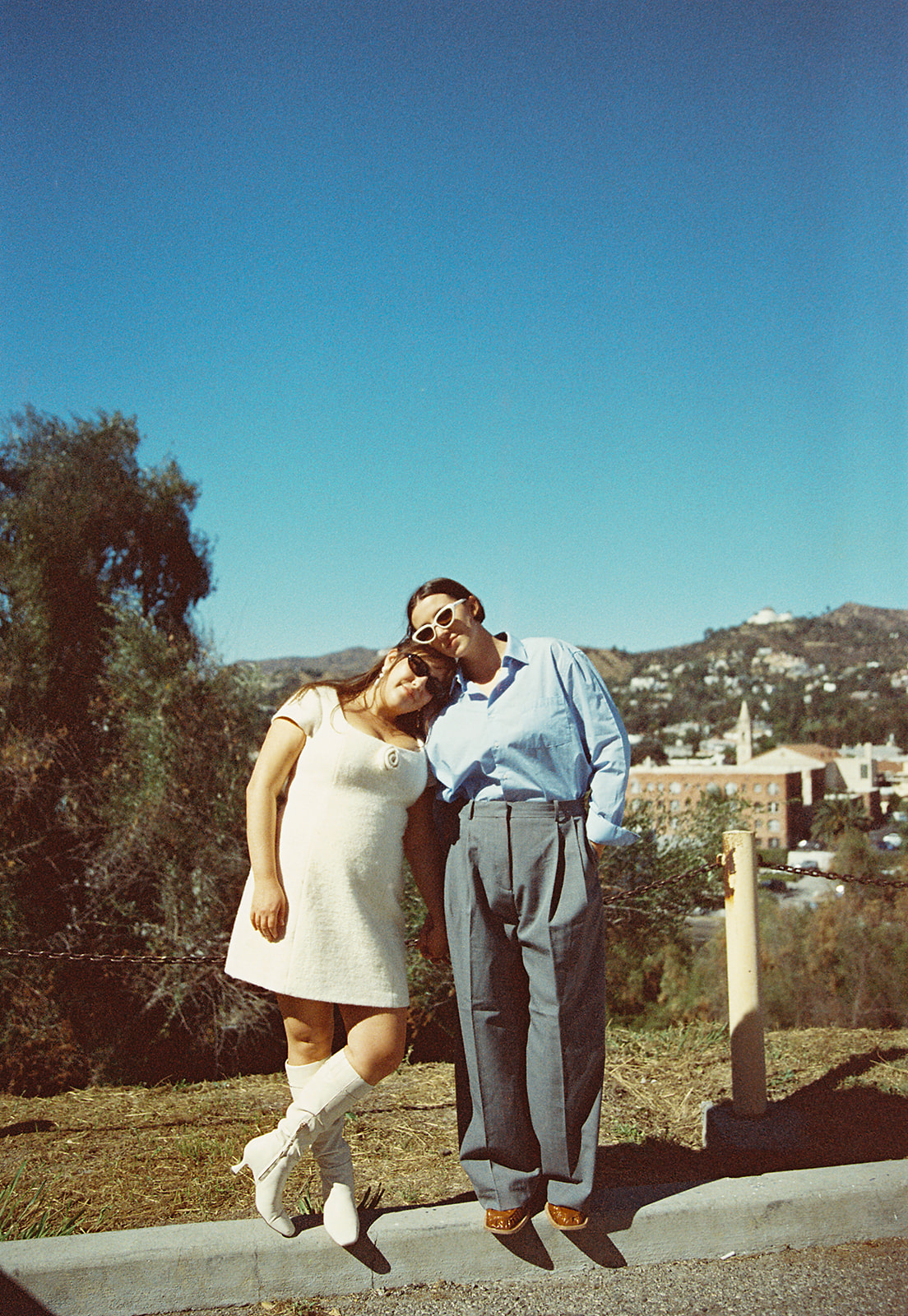 A couple holding hands on a sunny day with a cityscape in the background. The man is wearing a blue shirt and gray pants, and the woman is in a white dress and boots.