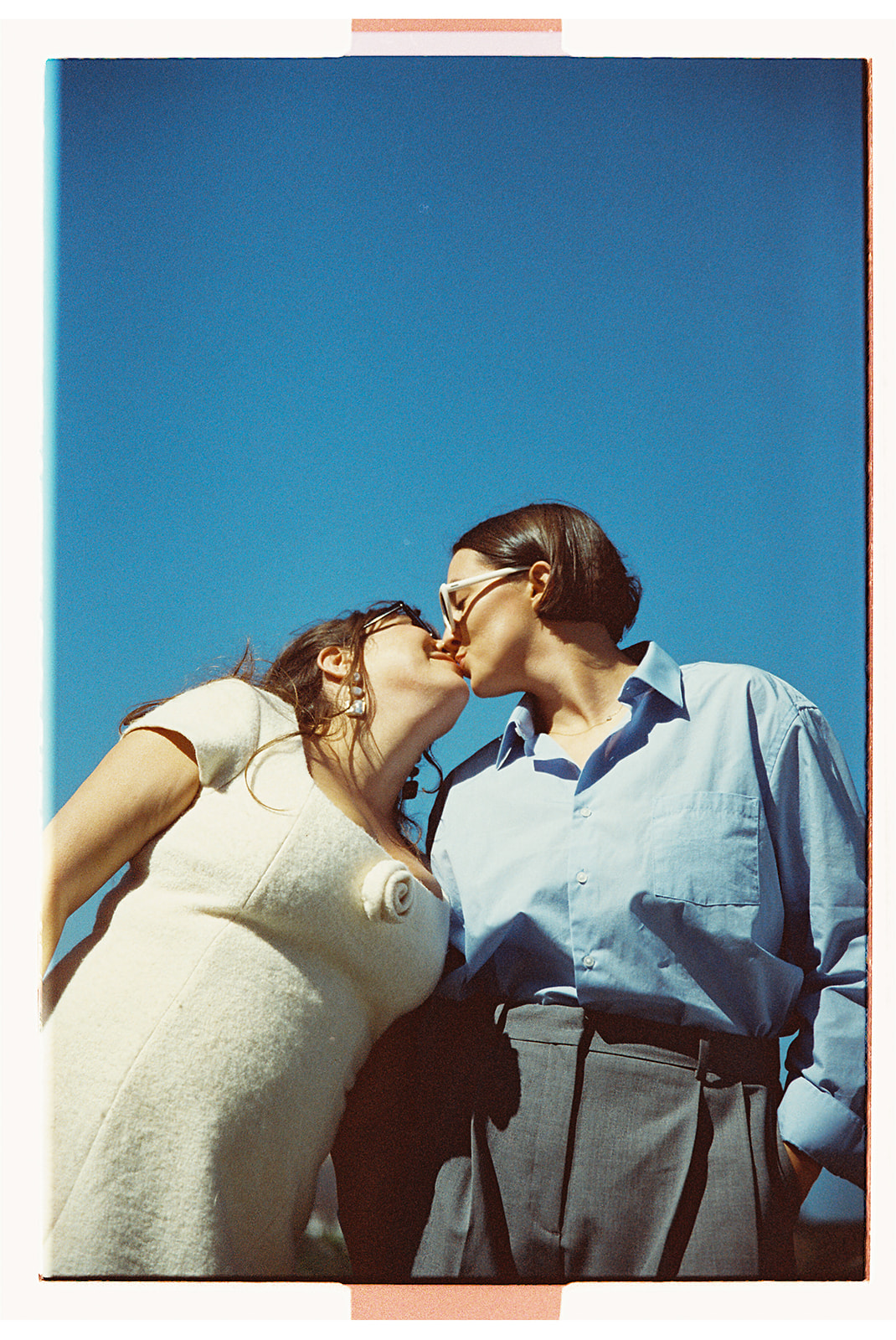 A couple holding hands on a sunny day with a cityscape in the background. The man is wearing a blue shirt and gray pants, and the woman is in a white dress and boots.