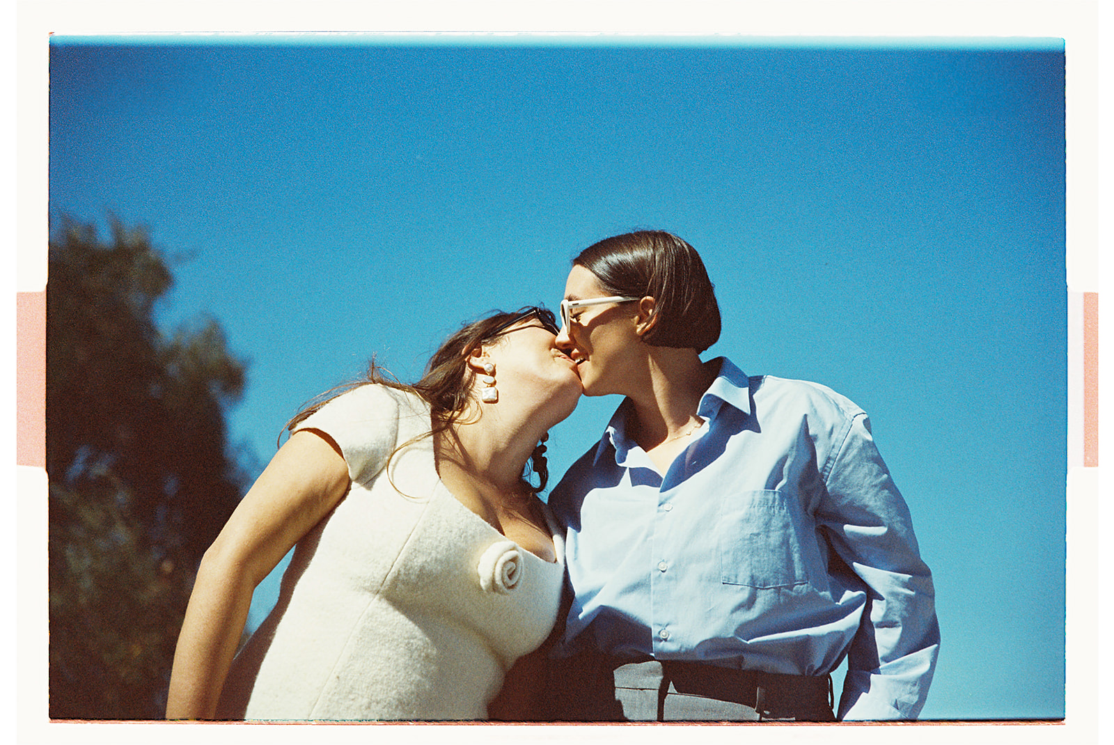 Two people are kissing against a clear blue sky. One is wearing a light-colored top, and the other is in a blue shirt.