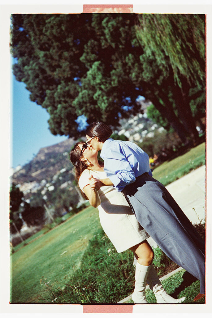 A couple shares a kiss outdoors on a sunny day, surrounded by greenery and trees.