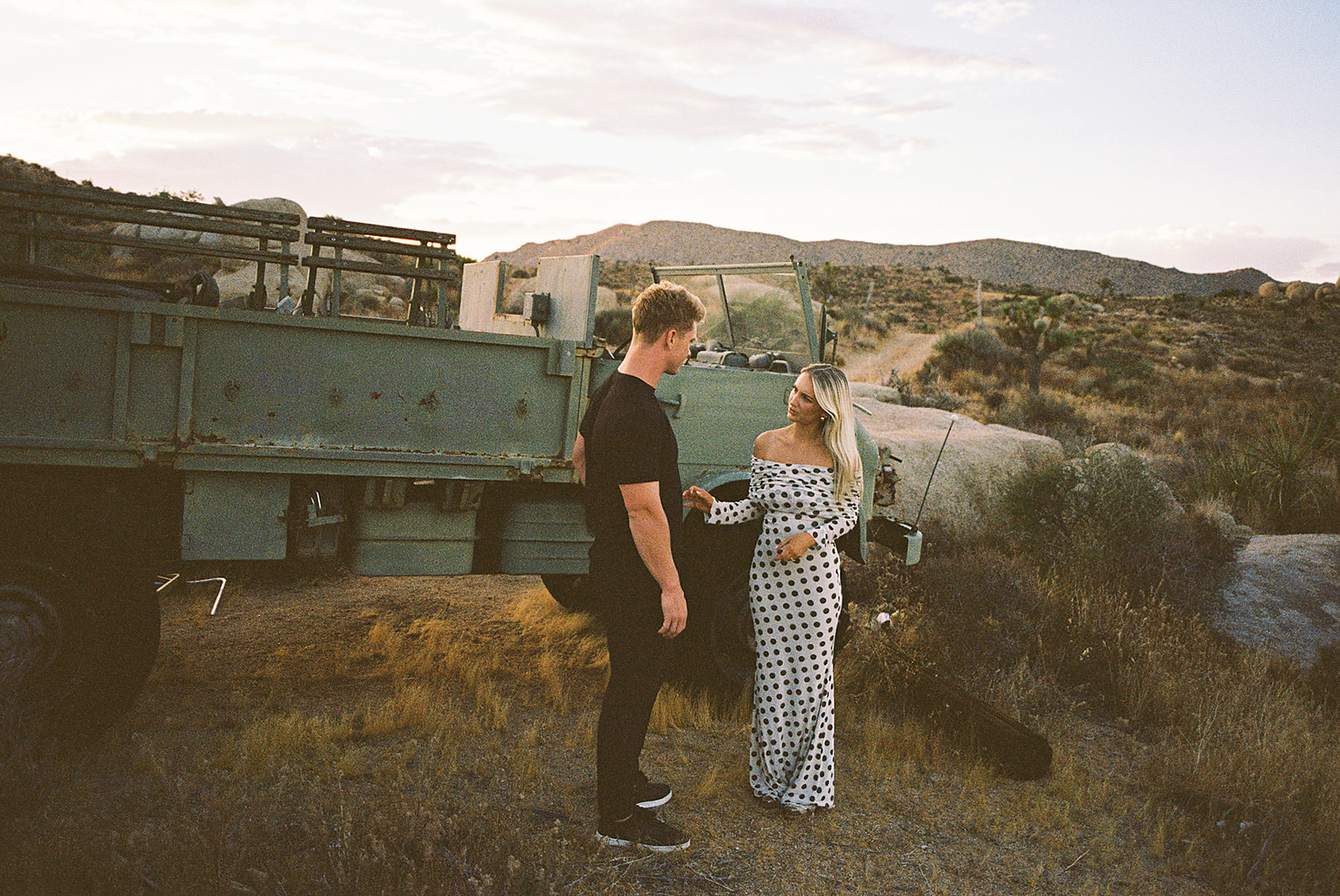 A couple walks hand in hand a dirt path. The woman wears a polka dot dress, and the man wears a black shirt and pants.