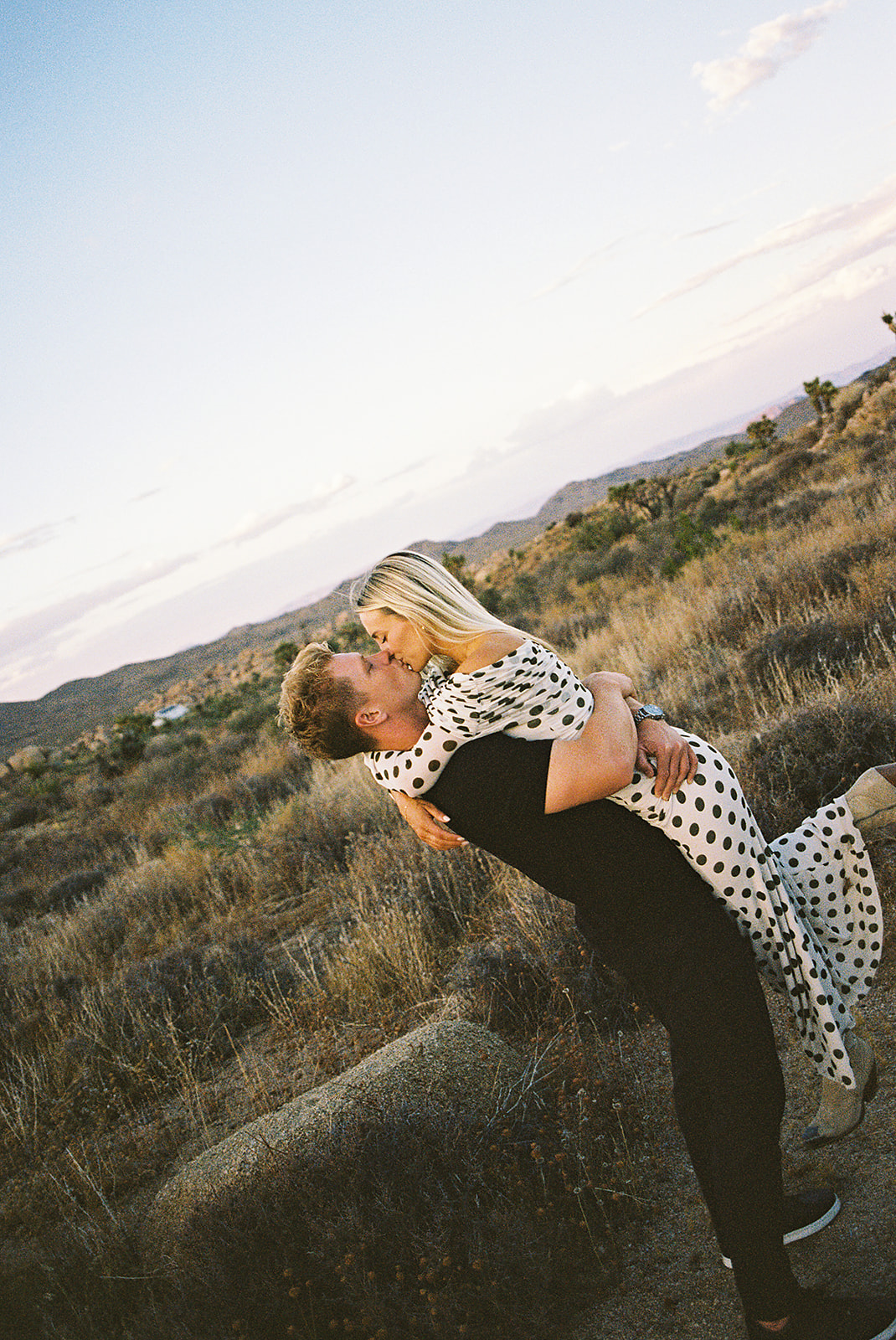 A couple walks hand in hand a dirt path. The woman wears a polka dot dress, and the man wears a black shirt and pants.