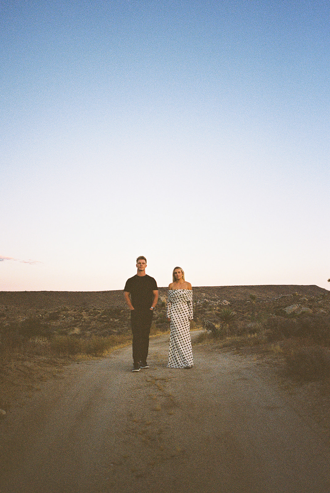 A couple walks hand in hand a dirt path. The woman wears a polka dot dress, and the man wears a black shirt and pants.