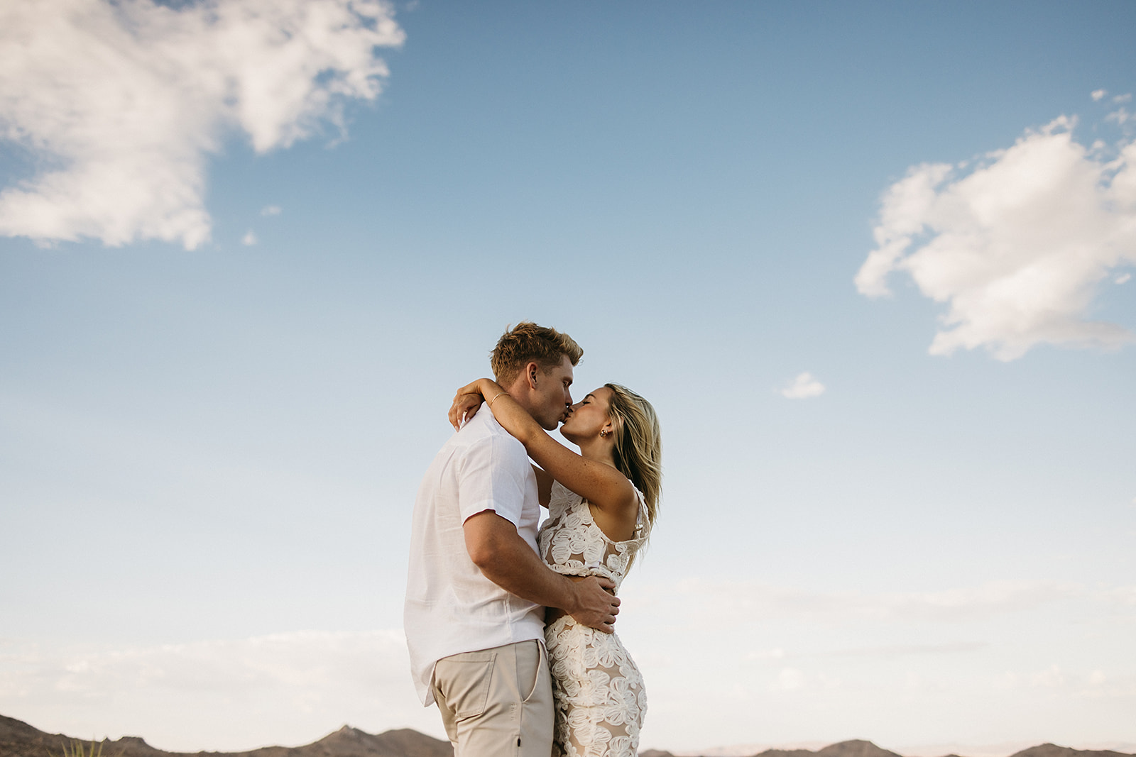 A couple holding hands walks through a desert landscape under a clear blue sky for their joshua tree engagement photos