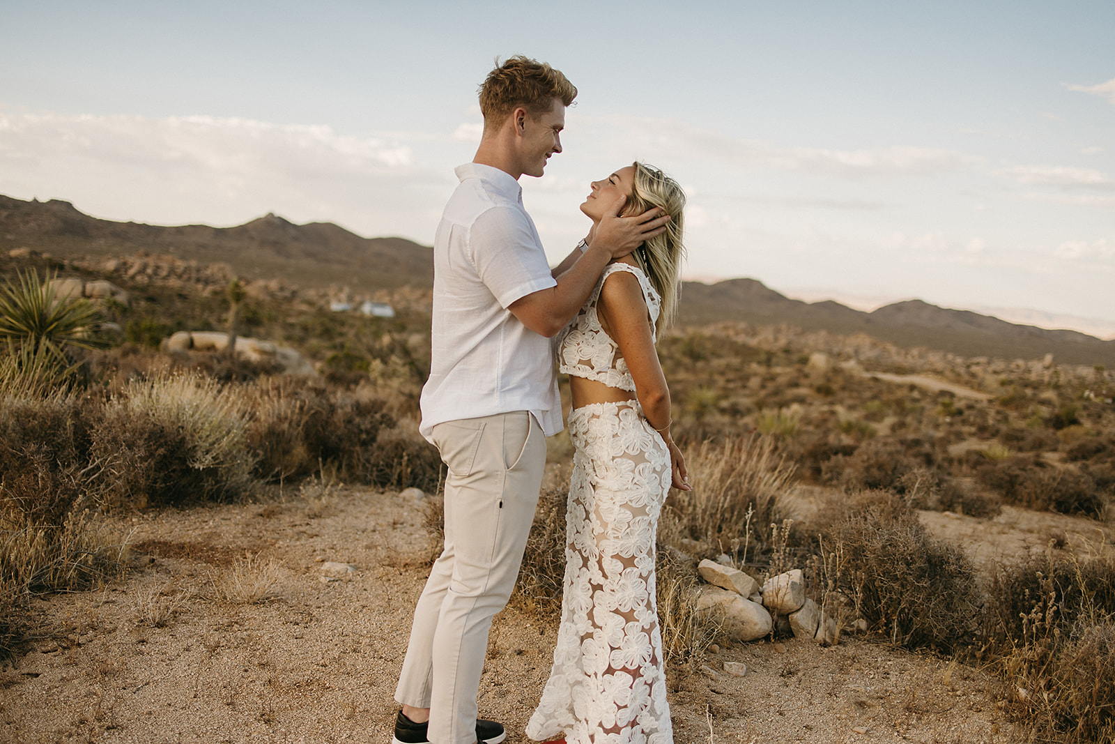 A couple holding hands walks through a desert landscape under a clear blue sky for their joshua tree engagement photos