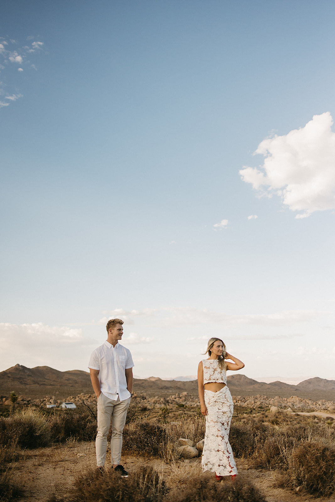 A couple holding hands walks through a desert landscape under a clear blue sky for their joshua tree engagement photos