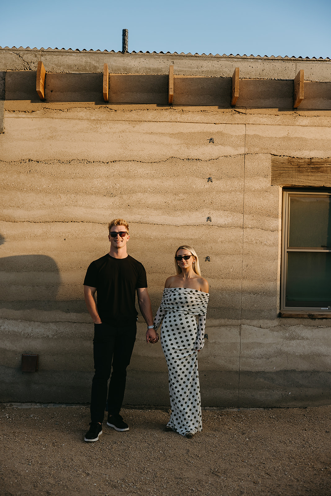 A person in a black outfit and sunglasses stands beside a person in a patterned dress and sunglasses, holding hands in front of a beige concrete wall.