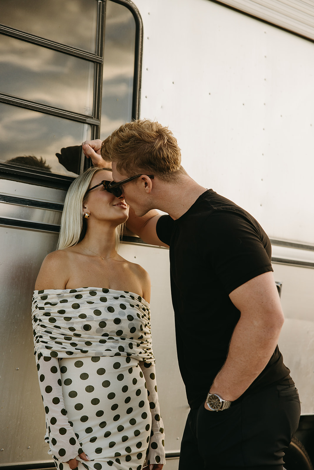 A couple leans in for a kiss in front of a shiny metal trailer. The woman wears a white dress with black polka dots, and the man is in a black shirt.