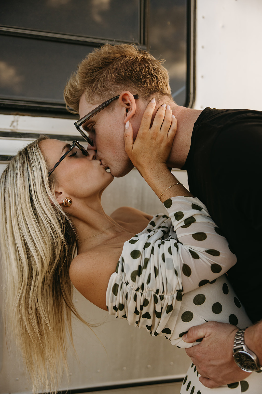 A couple leans in for a kiss in front of a shiny metal trailer. The woman wears a white dress with black polka dots, and the man is in a black shirt.