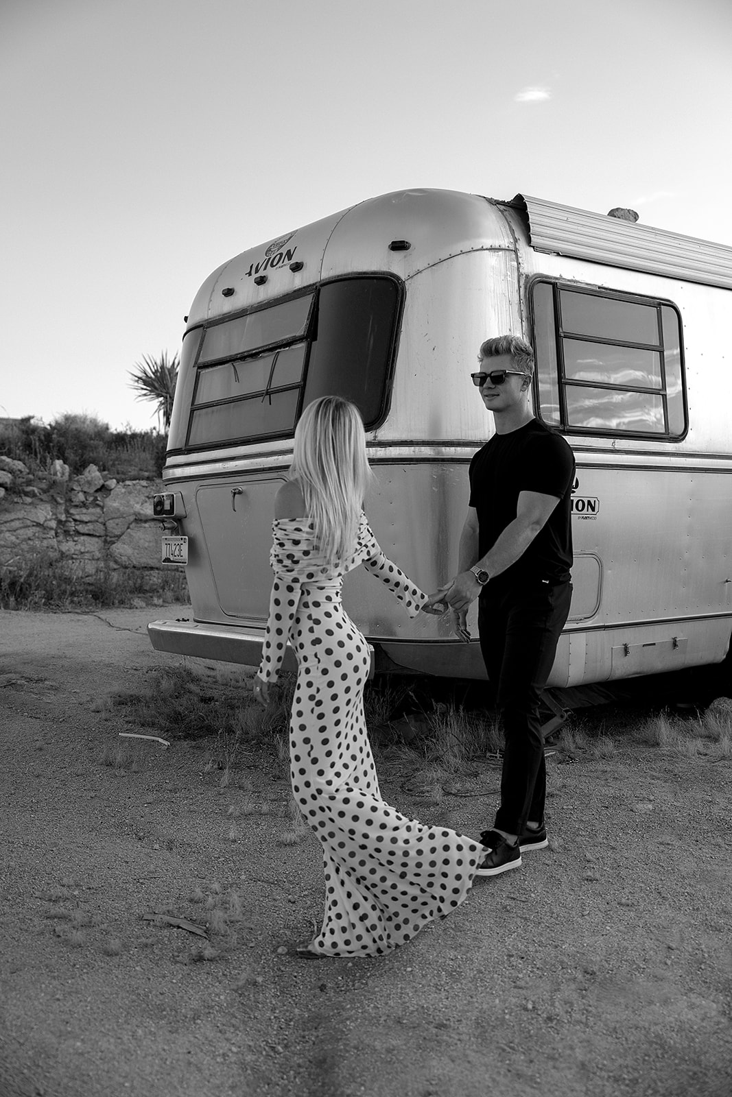 A couple walks hand in hand near a silver trailer on a dirt path. The woman wears a polka dot dress, and the man wears a black shirt and pants.