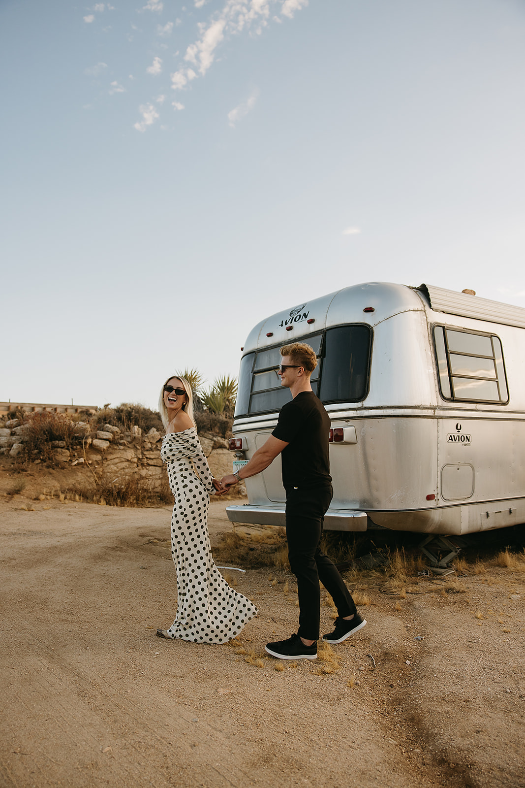 A couple walks hand in hand near a silver trailer on a dirt path. The woman wears a polka dot dress, and the man wears a black shirt and pants.