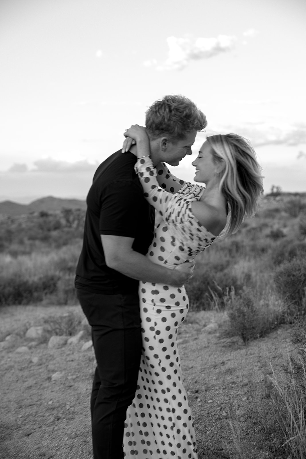 A couple walks hand in hand a dirt path. The woman wears a polka dot dress, and the man wears a black shirt and pants.