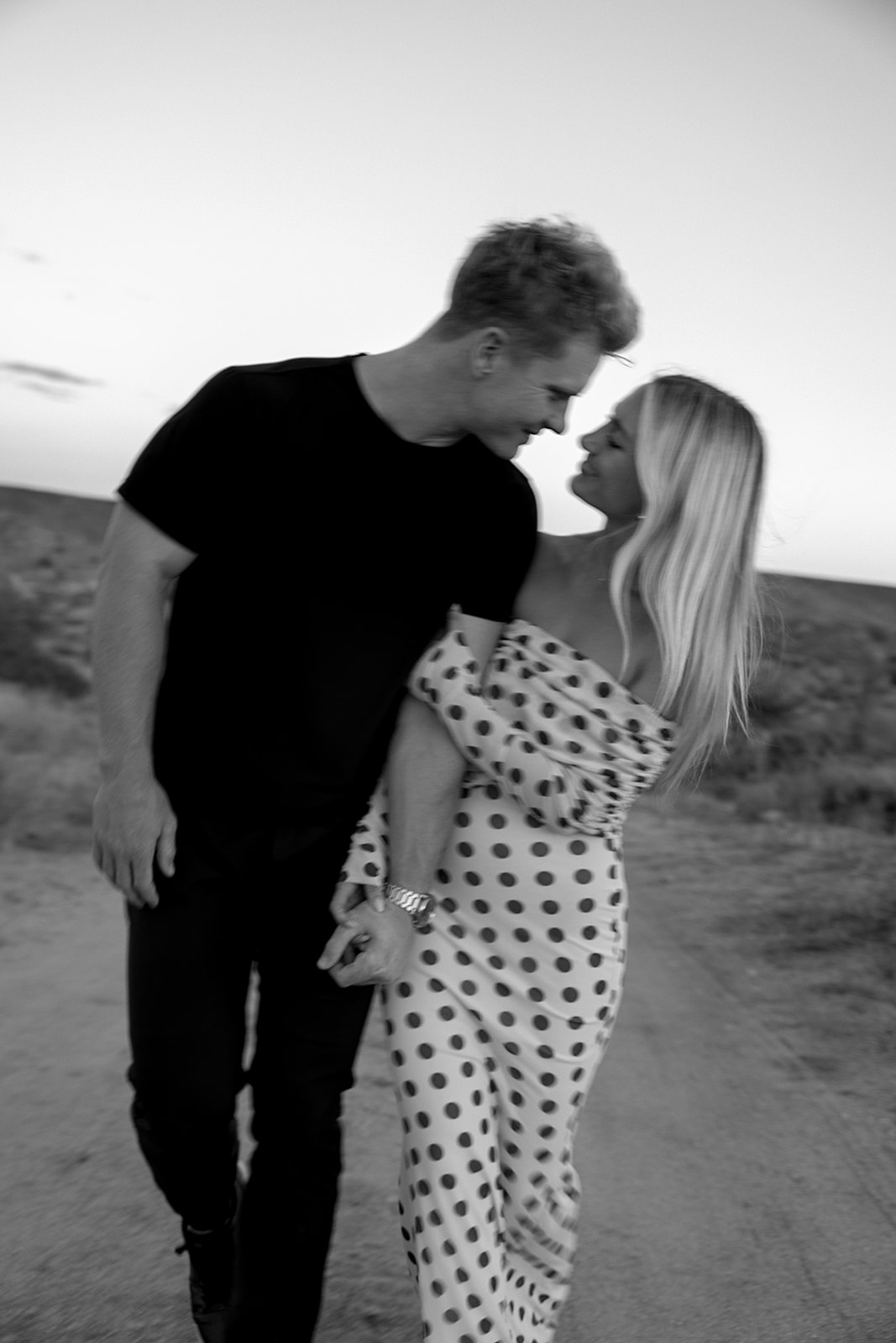 A couple walks hand in hand a dirt path. The woman wears a polka dot dress, and the man wears a black shirt and pants.