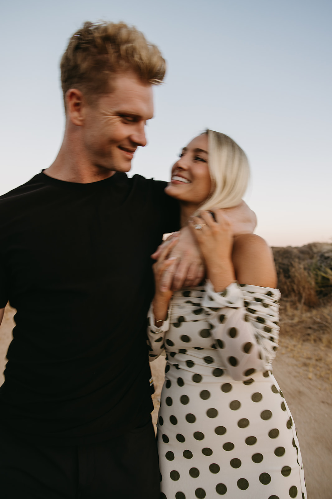 A couple walks hand in hand a dirt path. The woman wears a polka dot dress, and the man wears a black shirt and pants.