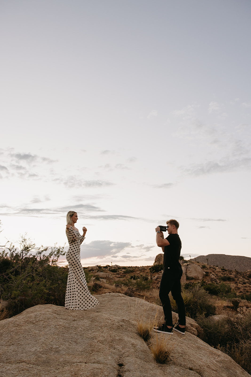 A couple walks hand in hand a dirt path. The woman wears a polka dot dress, and the man wears a black shirt and pants.