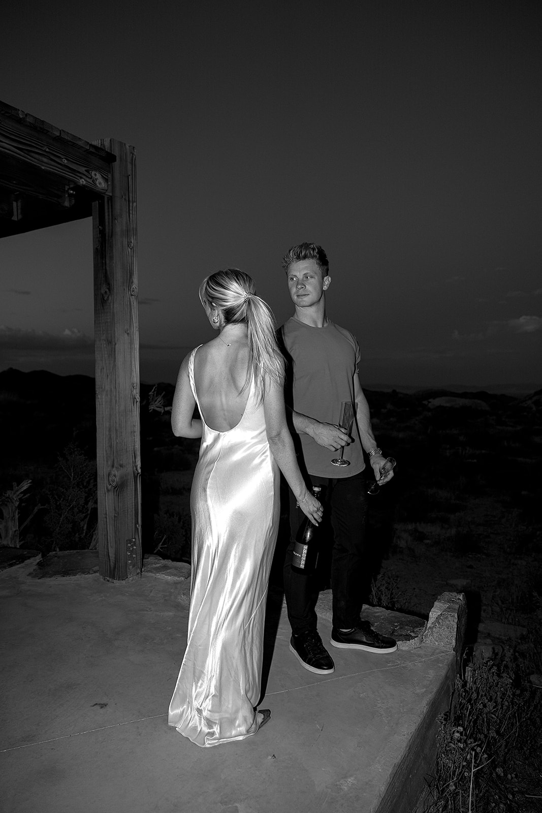 A person in a long dress stands next to a person in casual clothes holding drinks, on an outdoor patio at dusk for their joshua tree engagement photos