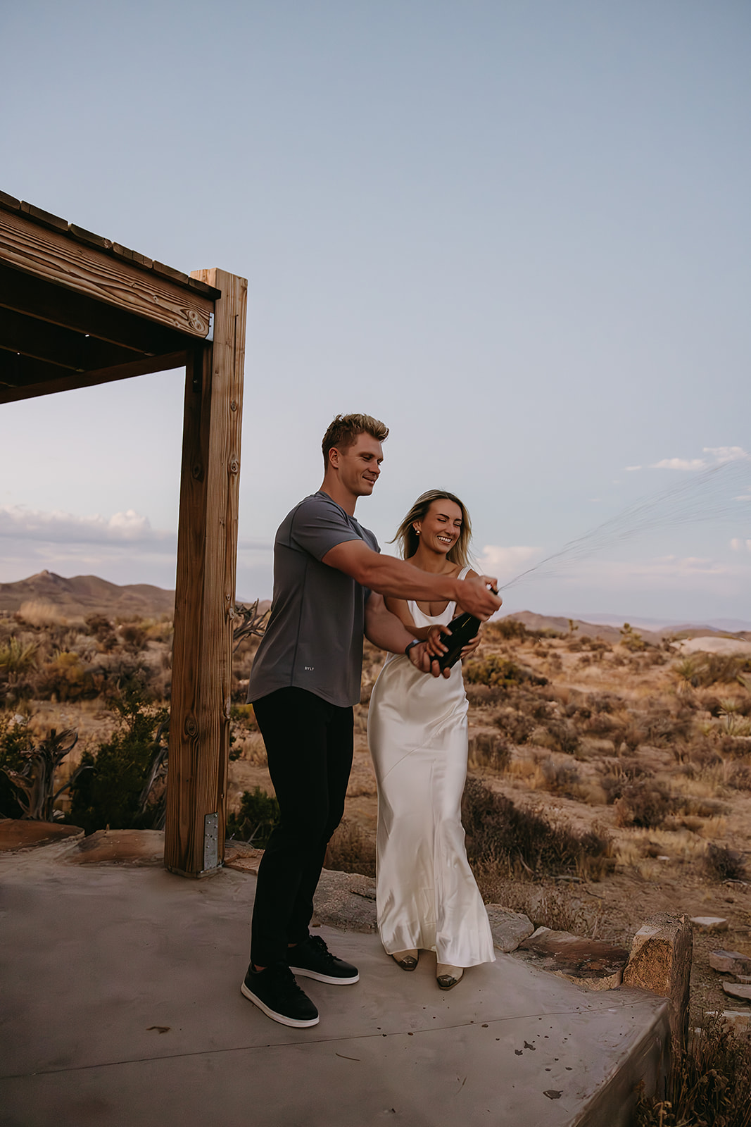 A man and woman stand on a concrete surface in a desert setting, joyfully spraying champagne.