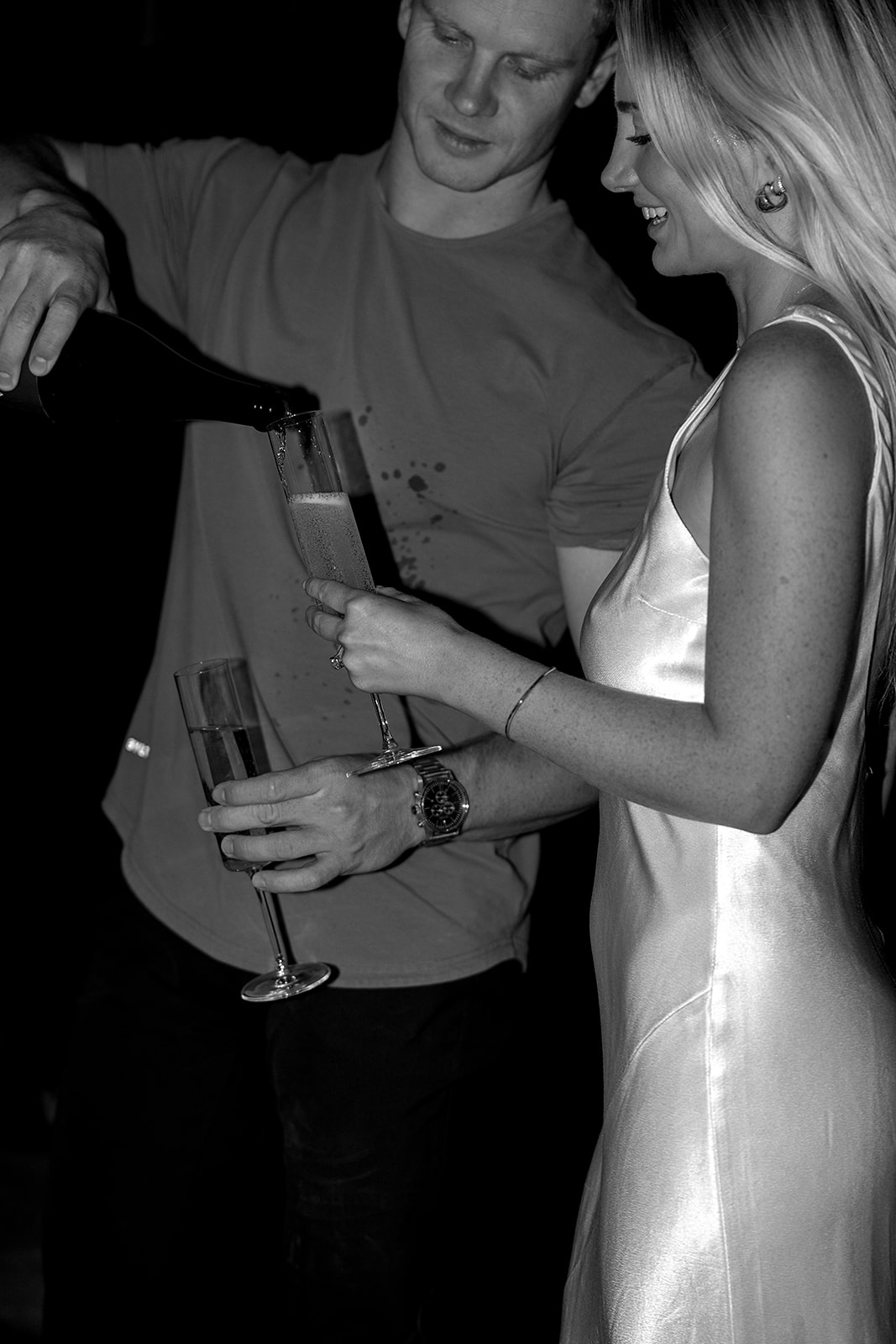 A person in a long dress stands next to a person in casual clothes holding drinks, on an outdoor patio at dusk for their joshua tree engagement photos