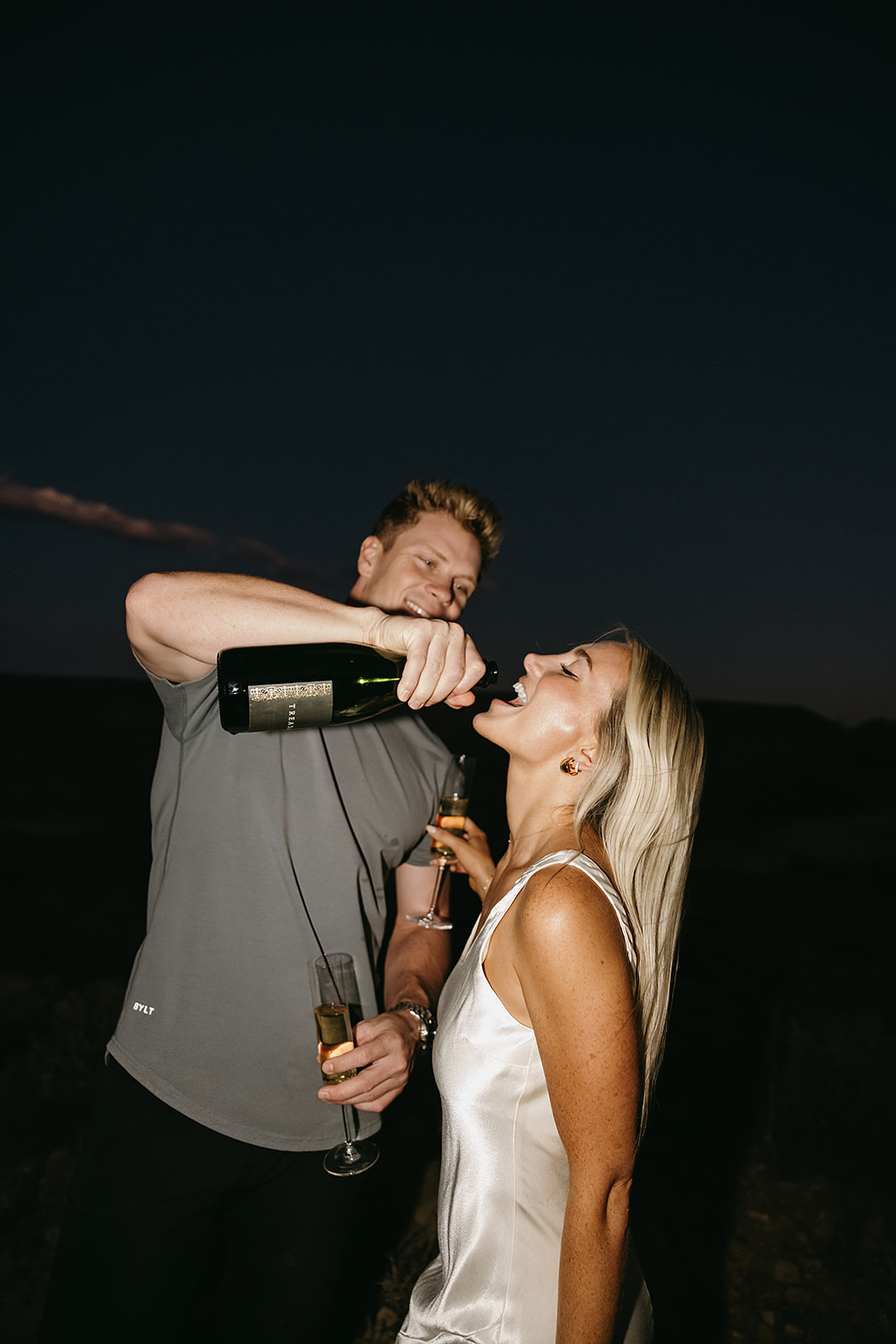 A man pours champagne into a woman's mouth outdoors at night. Both hold champagne glasses.