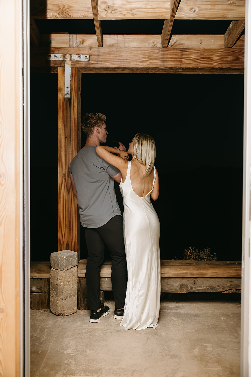 A couple stands on a wooden balcony, with the woman in a white dress, looking out into the night.