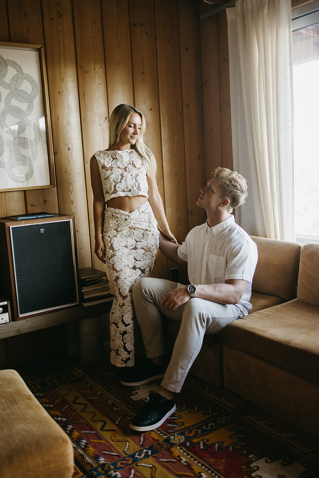 A couple sits closely on a sofa in a wood-paneled room. The man leans in to kiss the woman's cheek. A guitar rests against the wall, and a patterned art piece is visible in the background for their joshua tree engagement photos