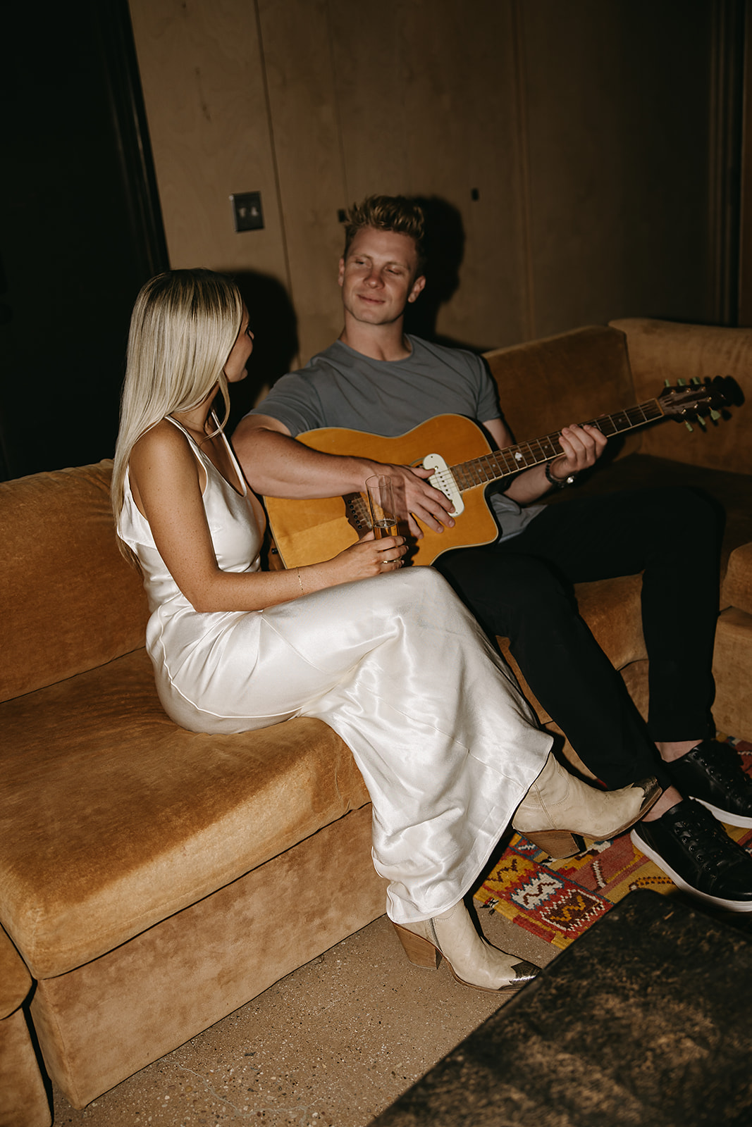 A person in a white dress sits on a couch holding a glass, while another person next to them plays a guitar for their joshua tree engagement photos