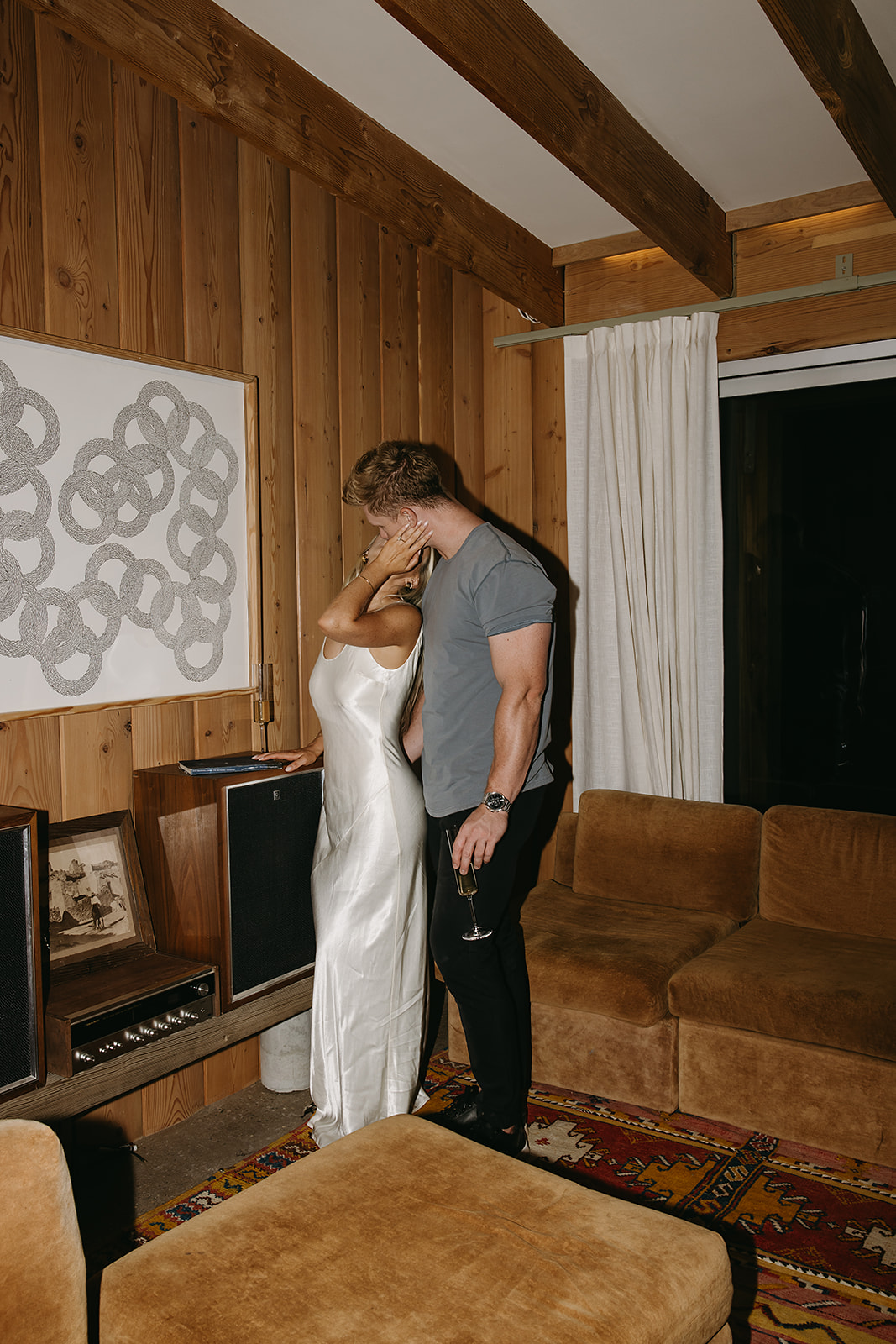 A person in a white dress sits on a couch holding a glass, while another person next to them plays a guitar for their joshua tree engagement photos