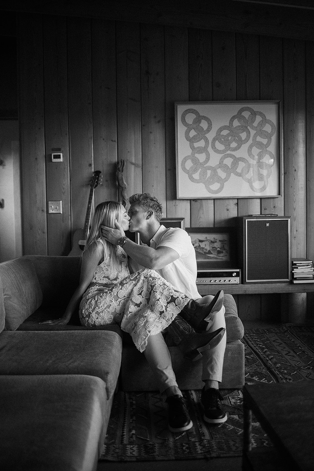 A couple sits closely on a sofa in a wood-paneled room. The man leans in to kiss the woman's cheek. A guitar rests against the wall, and a patterned art piece is visible in the background for their joshua tree engagement photos