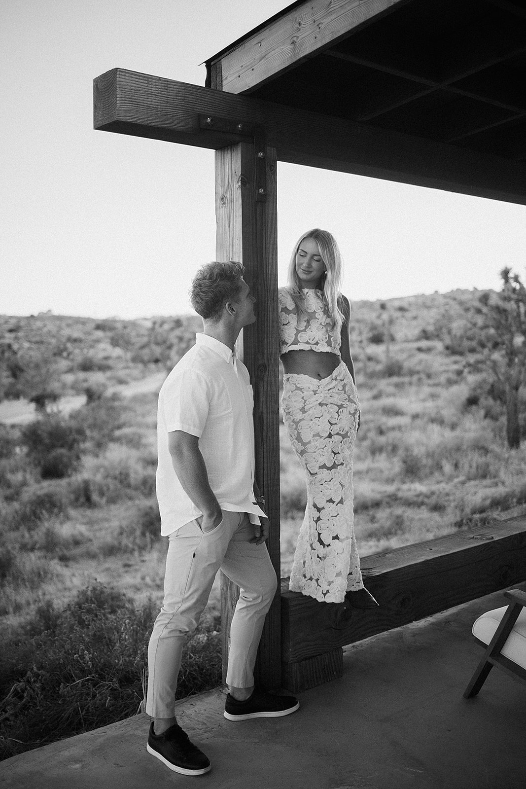 A man and woman face each other under a wooden structure in a desert landscape. The woman stands on a ledge, wearing a long skirt and crop top; the man wears a polo shirt and pants for their joshua tree engagement photos