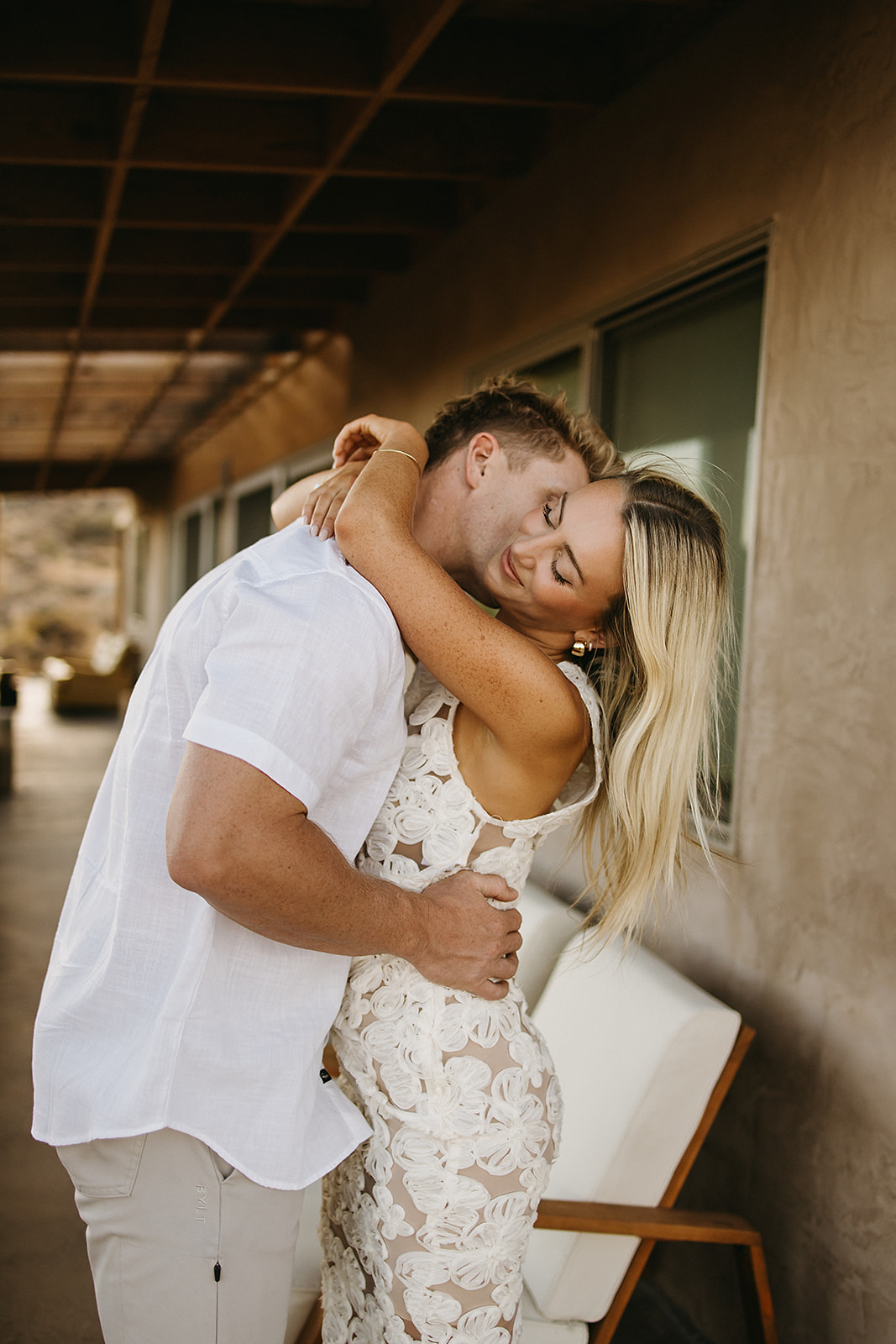 A man and woman face each other under a wooden structure in a desert landscape. wearing a long skirt and crop top; the man wears a polo shirt and pants for their joshua tree engagement photos