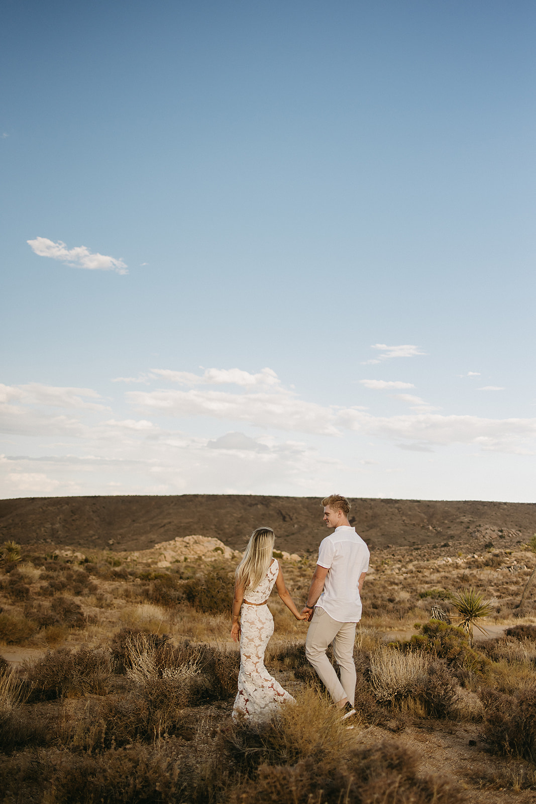 A couple holding hands walks through a desert landscape under a clear blue sky for their joshua tree engagement photos