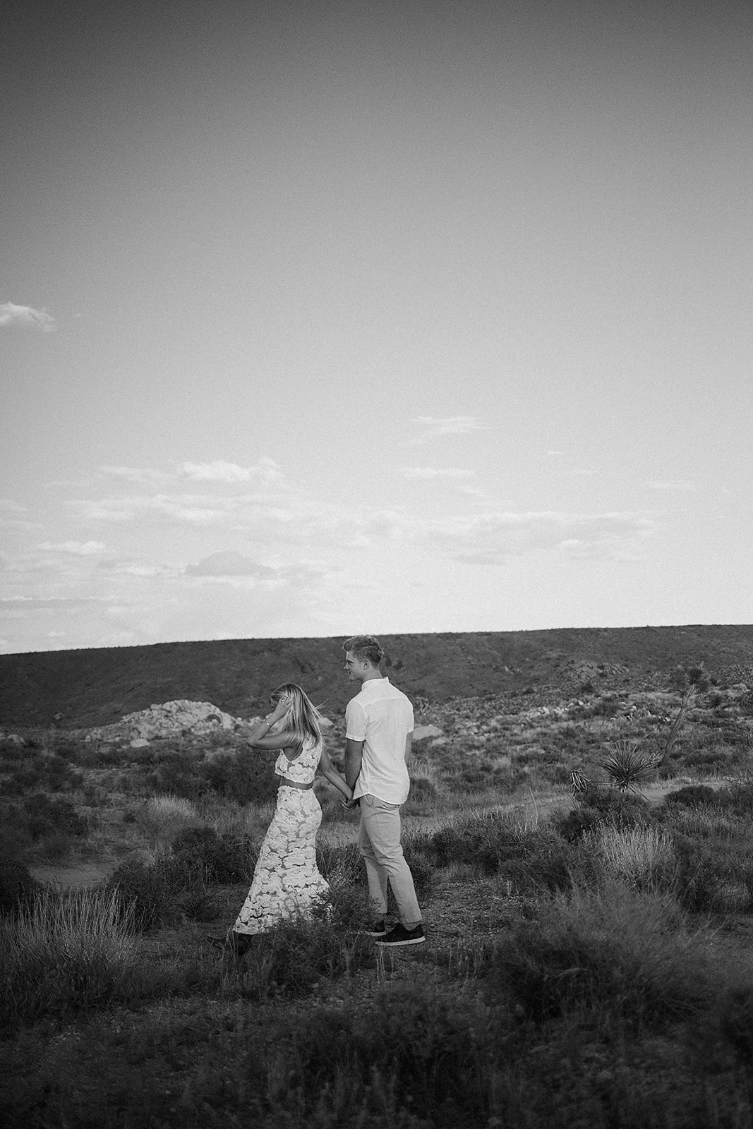 A couple holding hands walks through a desert landscape under a clear blue sky for their joshua tree engagement photos