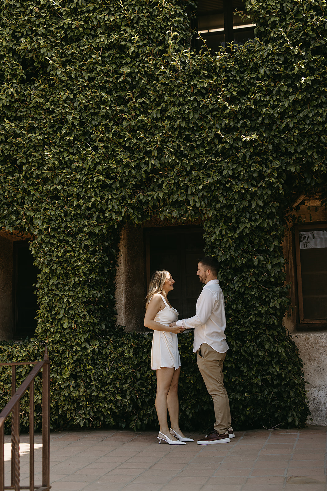 A couple stands close and embraces on a sunlit patio, with steps and a dark doorway in the background for their candid engagement photography 